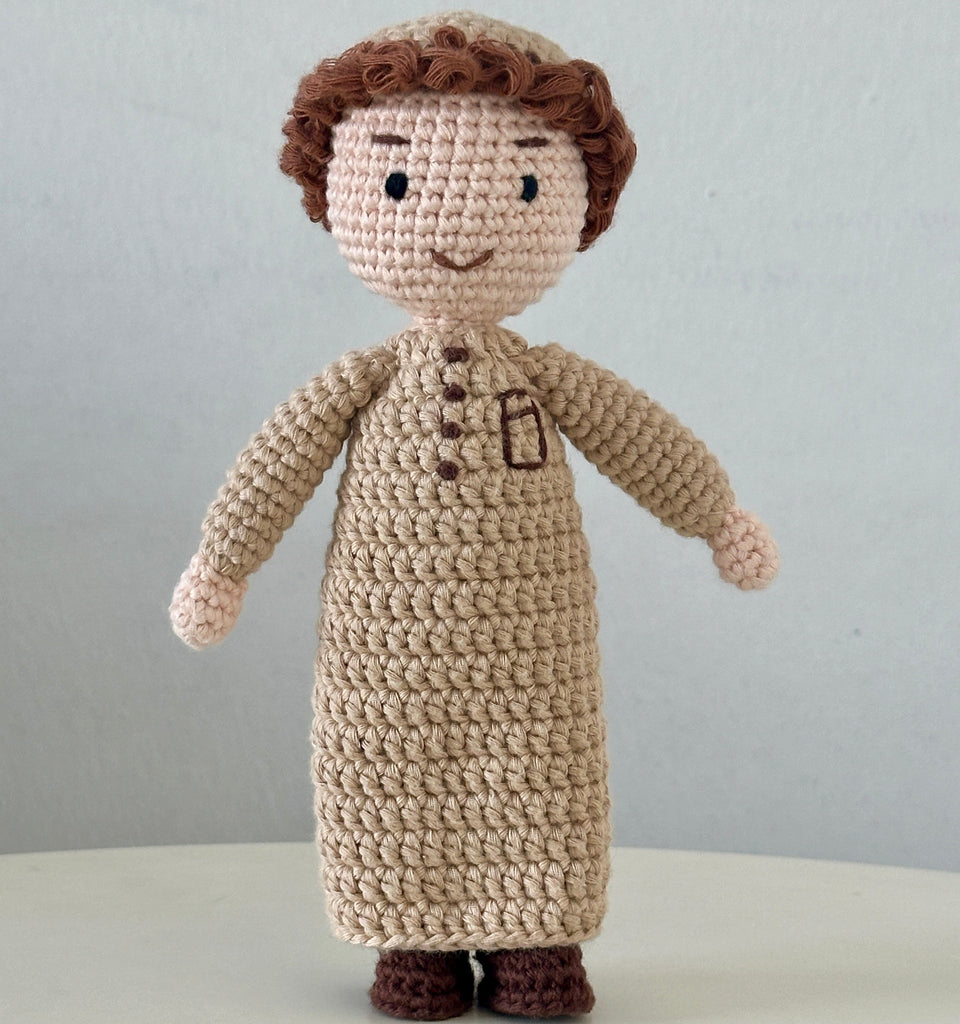 Crocheted doll with a brown hat and coat on a white surface with a gray background