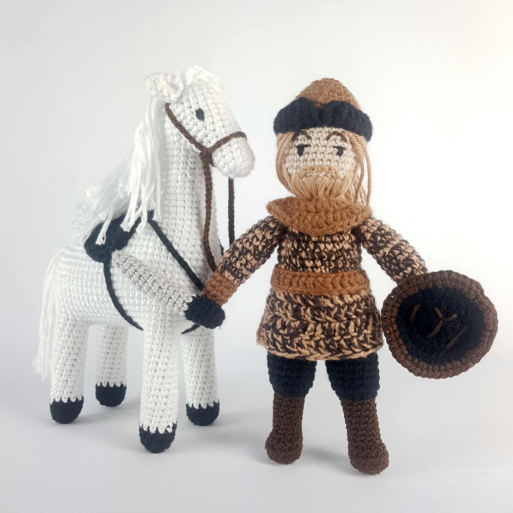alt="Handmade Ertugrul male hero crochet doll standing guard with long blond hair and beard, black and brown outfit an ottoman hat and a shield in his hand, other hand holding brown leash to beautiful white crochet horse with black saddle"
